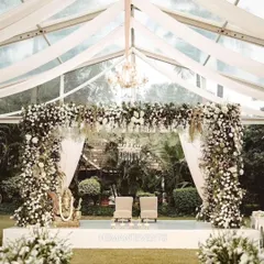 A beautiful wedding stage set inside a transparent tent, allowing for an outdoor feel with indoor comfort. The stage is heavily decorated with white flowers and features a Ganesha idol for blessings.