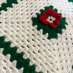 A close-up detail of the crochet cushion cover, highlighting the texture of the granny square stitch and the central red flower.