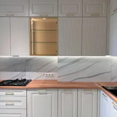 A classic white kitchen with a warm wooden countertop. The design features elegant moulded doors and a special crockery unit with a gold-finished interior and glass shelves for a beautiful display.