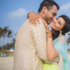 A close-up, candid moment of pure joy as the groom kisses the bride's cheek on the beach, her mehendi-adorned hands framing his face.