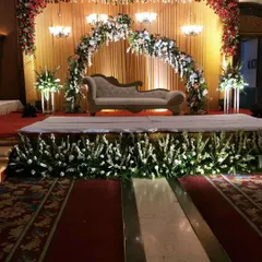 A sophisticated stage design with a floral arch in pastel shades. The setup includes a classic diwan sofa, elegant chandeliers, and a fully decorated stage front with white flowers.