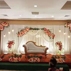 A grand stage decoration for a formal engagement or reception in a hall. The design features a wide white curtain backdrop, an arch of red and white flowers, and a classic white chaise lounge for elegant seating.