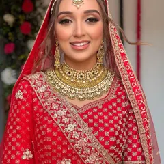 A beautiful bridal portrait. The hair is styled in a soft, face-framing updo with loose waves, complementing the delicate maang tikka and red lehenga for a classic bridal look.