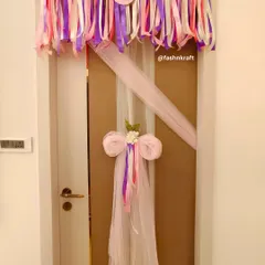 A playful and pretty decoration for a baby girl's room door. A colorful ribbon garland hangs above, while a large tulle bow with trailing ribbons adds a touch of whimsy and charm.