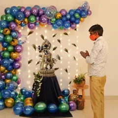 For a home puja or Janmashtami celebration, I can create a beautiful and sacred space. This setup features a balloon arch in peacock-inspired colors, a backdrop of fairy lights and feathers, and a decorated pedestal for the Krishna idol.