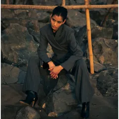 A full-length shot of the model seated on beach rocks. This pose feels both powerful and relaxed, showcasing the versatility of the styling in a natural setting.