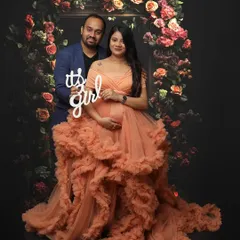 A joyful gender reveal moment captured within a maternity shoot. The couple is framed by a beautiful floral arch, and the mom-to-be wears a stunning ruffled peach gown, adding to the celebratory feel.