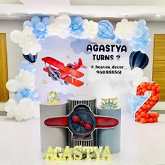 An adventurous airplane-themed second birthday party. The decor features a sky-blue backdrop with a vintage red airplane, a balloon garland resembling clouds, and a large mosaic number '2' filled with red balloons.