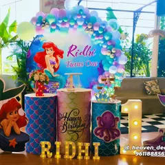 A vibrant Little Mermaid theme for a first birthday, featuring a circular backdrop with Ariel. The setup includes themed plinths with scale patterns, character cutouts, and a large light-up number '1'.