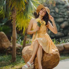 A sunny, cheerful lifestyle portrait. The model's yellow summer dress, straw hat, and the lush green background come together to create a warm and joyful outdoor image.