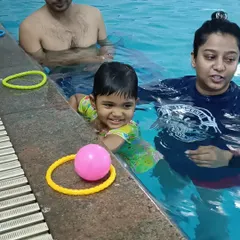 A smiling toddler plays with toys by the pool edge during an infant swim class, showing the fun and safe environment.