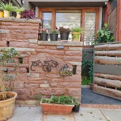 The entrance to a home named 'Samskruthi'. The rough-hewn stone compound wall and a gate made of unfinished wood logs create a rustic and earthy welcome, grounded in natural textures.