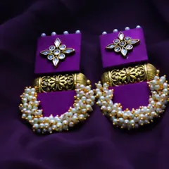 A detailed product shot of handmade purple and gold earrings against a matching purple fabric, emphasizing their color and intricate beadwork.