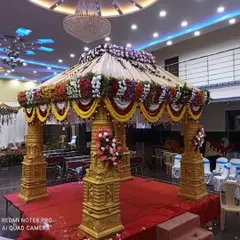 A classic four-pillared wedding mandap decorated with vibrant yellow and red flower garlands, set under a beautiful chandelier in a convention hall.