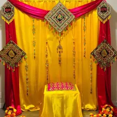 This compact Haldi setup is perfect for smaller indoor spaces. It combines a rich yellow backdrop with contrasting pink drapes and decorated with traditional embroidered hangings for a complete and festive look.
