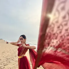 A candid moment during a beach photoshoot. The flowing saree against the sea breeze creates a beautiful, organic movement that complements the dance mudra.