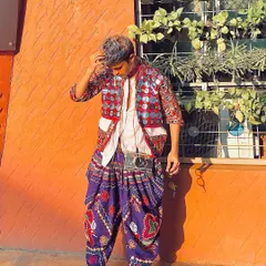 A cool, casual pose against a bright orange wall. This outfit features a mirror-work jacket over a simple white shirt and purple dhoti pants, a great way to style a modern Garba look.