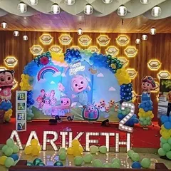 A full view of a Cocomelon stage decoration for a second birthday. The setup includes a large backdrop, balloon columns, character posters, and a light-up number '2' in a banquet hall setting.