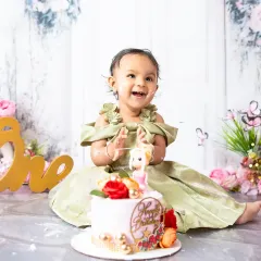 Pure happiness captured during a first birthday cake smash session. Dressed in a beautiful gown amidst a floral setup, this little one's joyful expression is what these celebratory milestone photoshoots are all about.
