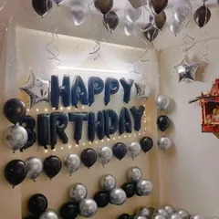 A classic home birthday decoration with black and silver balloons on the ceiling, "Happy Birthday" foil letters, and fairy lights.
