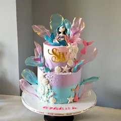 A dreamy, two-tiered mermaid cake in pastel shades of purple and blue. This design features a hand-sculpted mermaid, edible seashells, and shimmering rice paper sails to create an ethereal under-the-sea effect.
