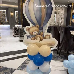 A close-up of a decorative element from the Mundan ceremony. This standalone piece features a cute teddy bear in a hot air balloon cutout, placed on a column of blue, white, and beige balloons.