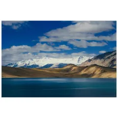 The stunning landscape of Tso Moriri in Ladakh, with snow-capped peaks reflecting in the deep blue waters under a partly cloudy sky.