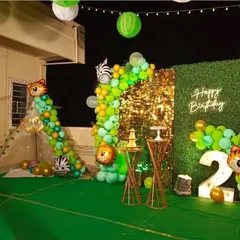 A wide view of an outdoor terrace jungle theme party for a second birthday. The setup includes a gold sequin and green wall backdrop, a light-up number '2', and a balloon garland that extends to a small teepee tent.