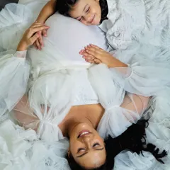 An artistic overhead shot capturing the sweet bond between mother and daughter. She rests her head peacefully on the baby bump, surrounded by the soft tulle of the maternity gown.