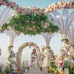 A beautiful shot of the couple under a spectacular floral mandap, with unique petal-like structures and a lush canopy of greenery and pink flowers against the open sky.