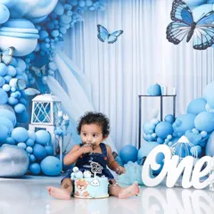 The moment before the smash. This photo shows little Naksha with his first birthday cake, set against an elaborate blue balloon and butterfly backdrop. The details of the setup are just as important as the action.