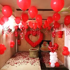 A complete room transformation designed for a big romantic gesture. The ceiling is covered with red balloons, a large heart made of smaller balloons serves as the centerpiece, and the floor is blanketed in red rose petals.