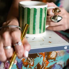 A detailed close-up of products in use for Patrika Gift Shop. This shot focuses on the hands holding a pen and a beautifully designed mug, highlighting the tactile experience of the brand's items.