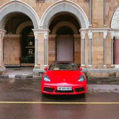 A bright red Porsche 718 Boxster parked in front of the ornate arches of a heritage building in South Bombay. The contrast between the modern sports car and the classic architecture is striking.