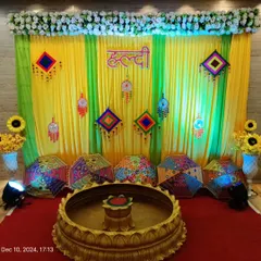 This Haldi decor features a vibrant backdrop with yellow and green curtains, decorated with colorful Rajasthani hangings and umbrellas. The setup includes a traditional urli for the ceremony.