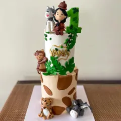 A jungle-themed two-tier cake for a first birthday celebration. The design features a giraffe print bottom tier, a vine-covered top tier, and adorable handmade fondant animals like a monkey, leopard, donkey, and a husky.
