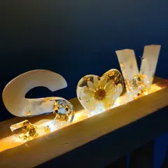 A custom resin name stand with embedded LED lights. The letters are filled with gold flakes and a single preserved daisy, creating a warm and personalized lamp for a desk or nightstand.