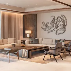 This living room at DLF Camellias showcases a blend of contemporary design with Indian elements. A neutral-toned sofa is set against a wall with textured paneling, while a large metal wall sculpture adds a dynamic focal point. The custom coffee table features an intricately carved metal top, reflecting our commitment to bespoke details.