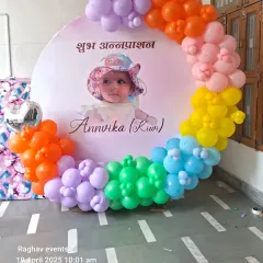 A colorful decoration for an Annaprashan ceremony. This setup features a custom round backdrop with "Shubh Annaprashan" written in Hindi and a photo of the baby, framed by a rainbow balloon garland.