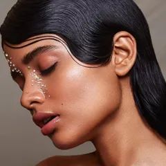 An artistic portrait featuring a model with a sleek, wet-look hairstyle and delicate rhinestone details near the eye. This image showcases precision in both hairstyling and makeup application.