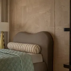 A detail of another bedroom in the Aikaa villa, showcasing the custom-designed curved headboard and wood-paneled wall. The mix of textures, from the quilted bedspread to the striped bolster, adds warmth and interest.