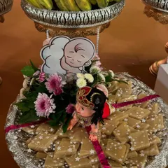 A sweet platter for a baby shower, decorated with a cute baby doll and a baby graphic. The tray is filled with kaju katli, making it a delicious and beautiful offering.