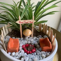 I created this miniature 'Home Sweet Home' garden to be a tiny escape on a desk. It features a spider plant, a quaint picket fence, and a whimsical campfire scene, all designed to bring a smile and a moment of peace to a busy workday.