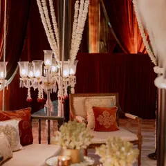 A close-up of a seating area from a royal-themed reception. The decor features elegant candelabras draped with jasmine, set against a backdrop of rich, red velvet curtains.