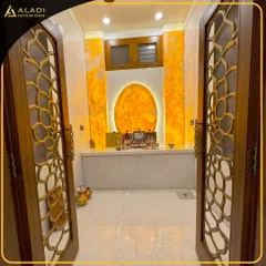 A view into a stunning puja room through intricately designed doors with a floral metal jali pattern. The back wall glows with a backlit onyx panel shaped like the sun, creating a divine ambiance.