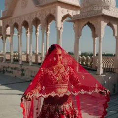 The bride playfully peeks through her vibrant red dupatta, her smile radiant against the backdrop of a royal Rajasthani palace.