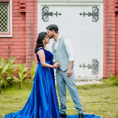 A tender moment as the partner kisses the mother-to-be's forehead. They stand in a beautiful blue gown before a charming building, creating a fairytale-like scene.