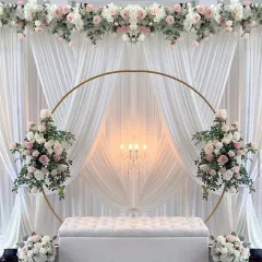 An absolutely breathtaking wedding or engagement stage. The setup features a large golden ring, elegant white draping, and beautiful arrangements of white and pink artificial flowers.