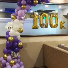 Celebrating a major milestone with a custom balloon arrangement. This organic half-arch in purple, white, and gold is paired with large '100K' foil balloons to mark the achievement, perfect for social media announcements or company events.