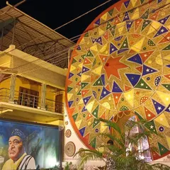 An exterior night view of the Durga Puja pandal, featuring a massive, colorful 'Japi' (traditional Assamese hat) and a mural of Bhupen Hazarika.